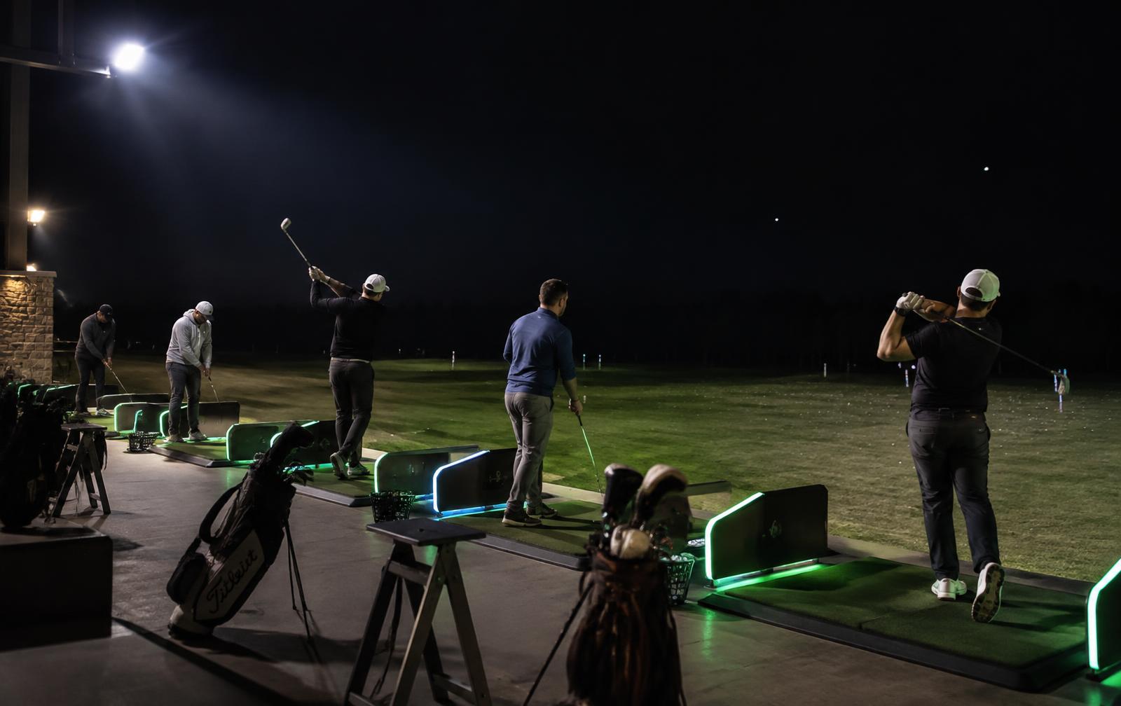 Jugadores practicando en el tee de práctica iluminado mientras el campo permanece a oscuras